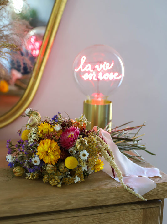 bouquet de mariée réalisé avec des fleurs séchées colorées. Le bouquet est posé sur un meuble à côté d'une grosse ampoule avec un message "la vie en rose"