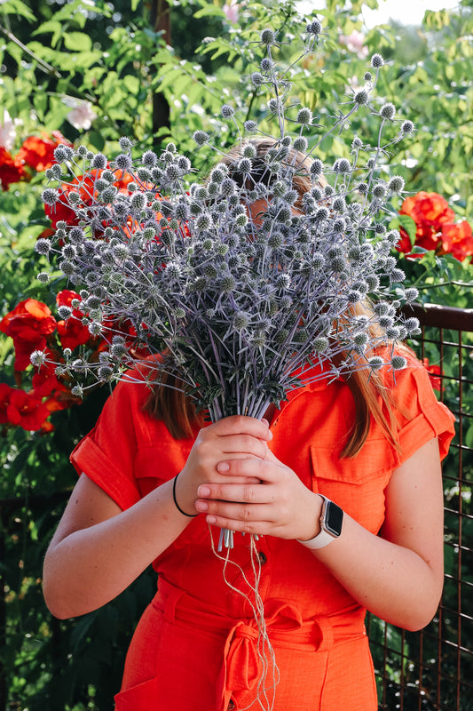 jeune femme habillées en orange qui tient un bouquet de fleurs bleu, des eryngium qui ressemblent à petits chardons bleus