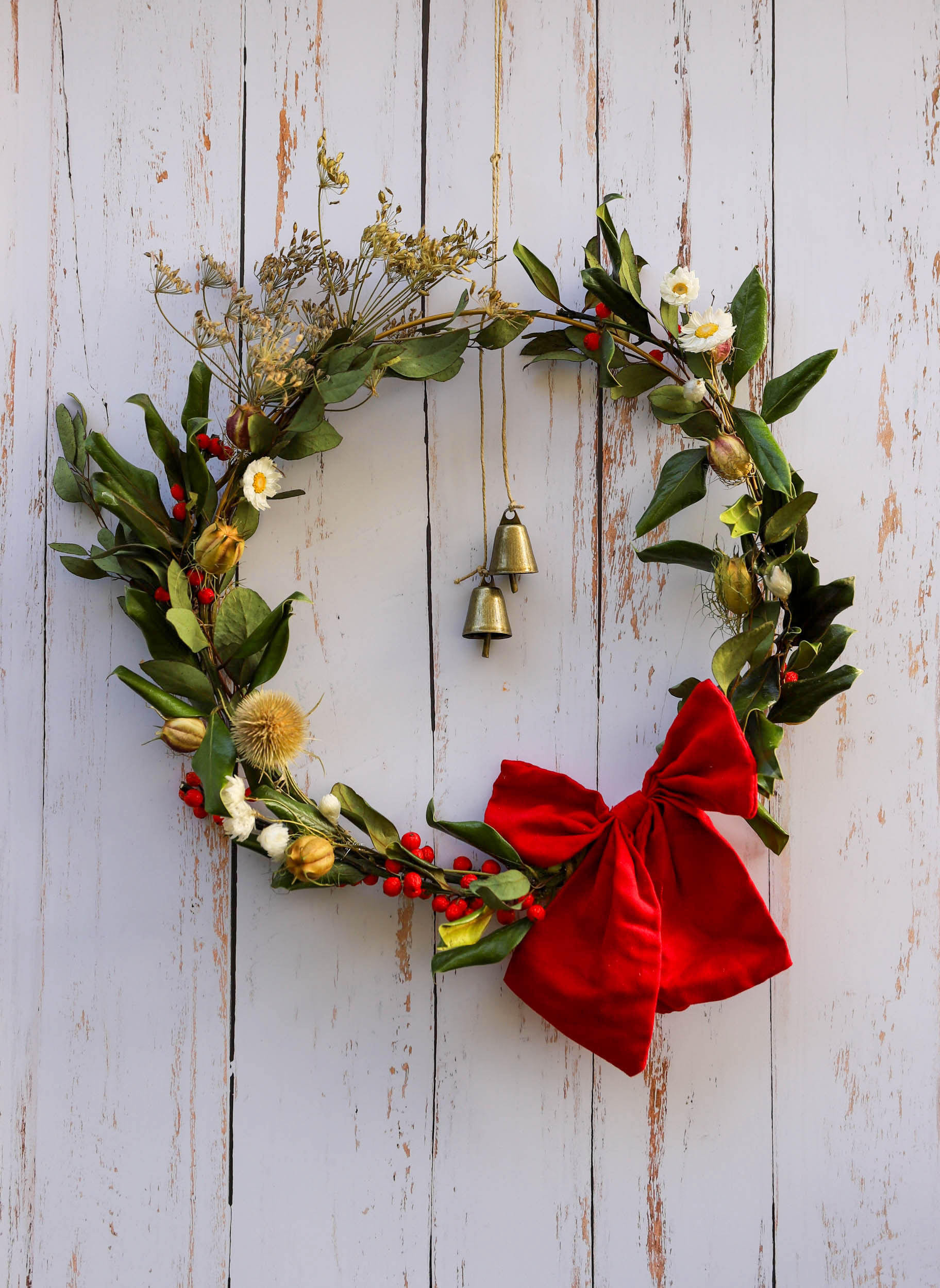 couronne de Noël avec des branches de houx, d'eucalyptus, chardon et fleurs blanches d'acroclinium. La couronne est ornée d'un gros noeud rouge et de deux clochettes dorées