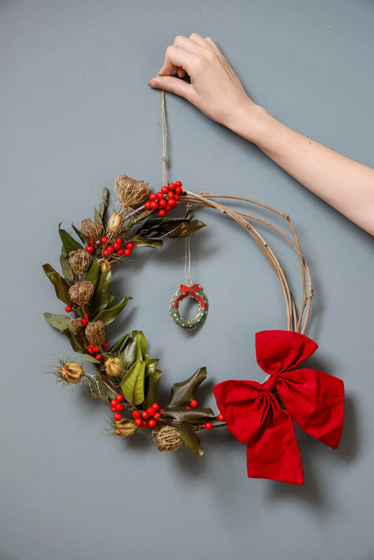 couronne de Noël réalisée avec des sarments de glycine, du houx avec des baies rouges, des gousses de nigelles, un gros noeud et une décoration représentant une couronne de Noël