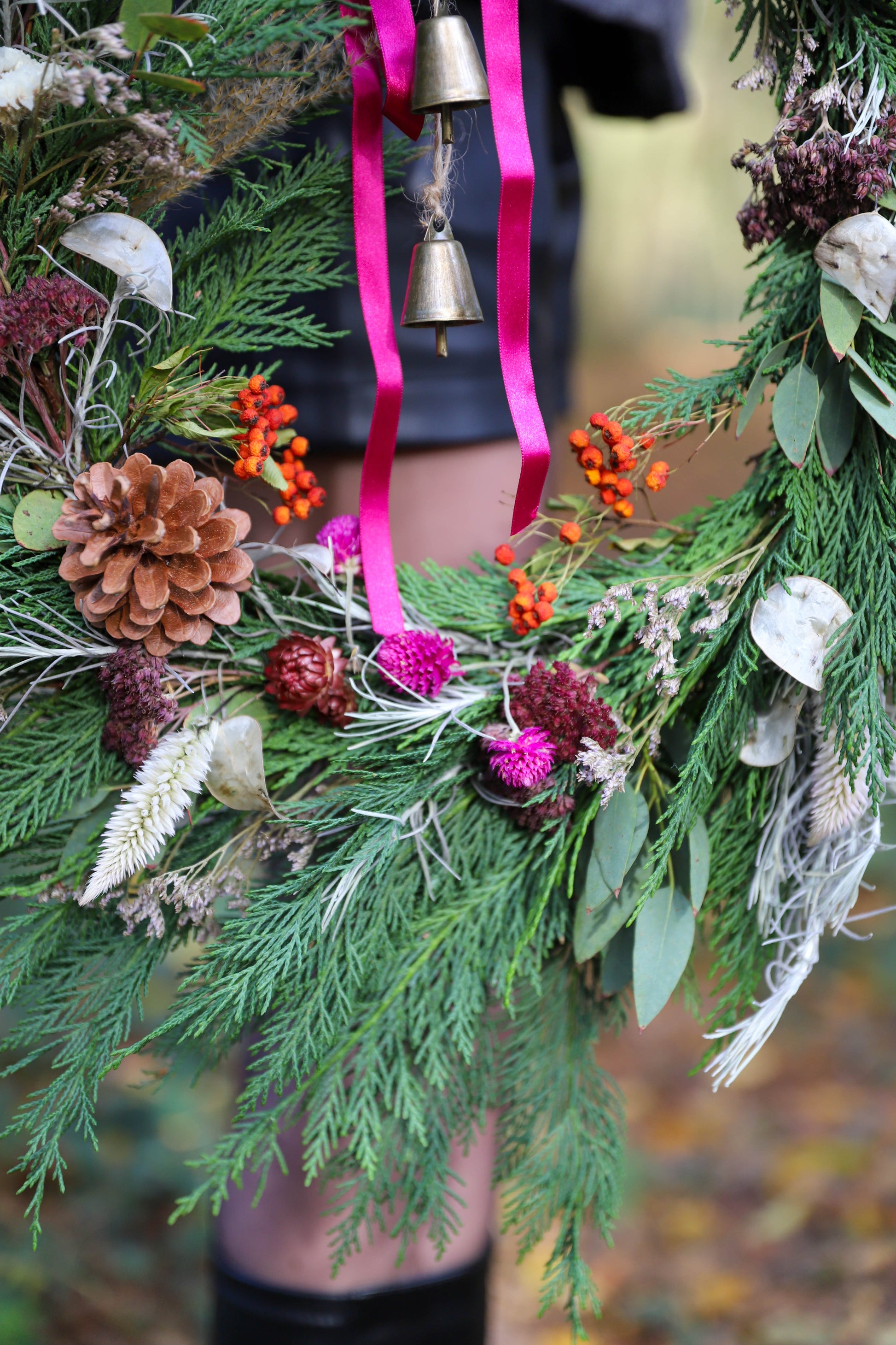 couronne de Noël française avec des baies rouges fraiches et des fleurs séchées colorées, il y a des cloches en métal et un ruban